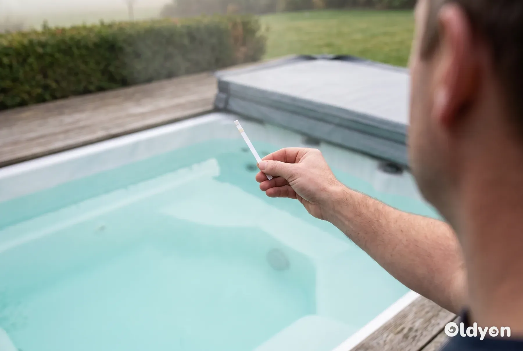 Hand seen from behind dipping a water test strip into a clear hot tub in a British garden on a soft, overcast day