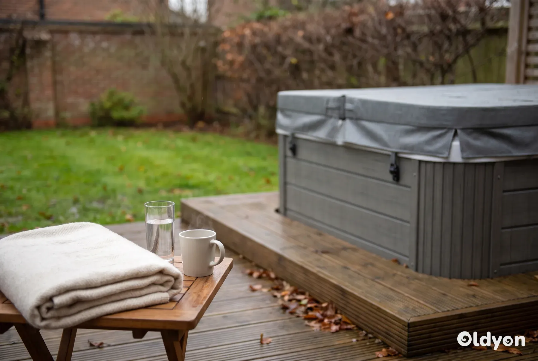 Covered hot tub in a damp British garden, with a folded blanket, a glass of water and a mug on a wooden table nearby