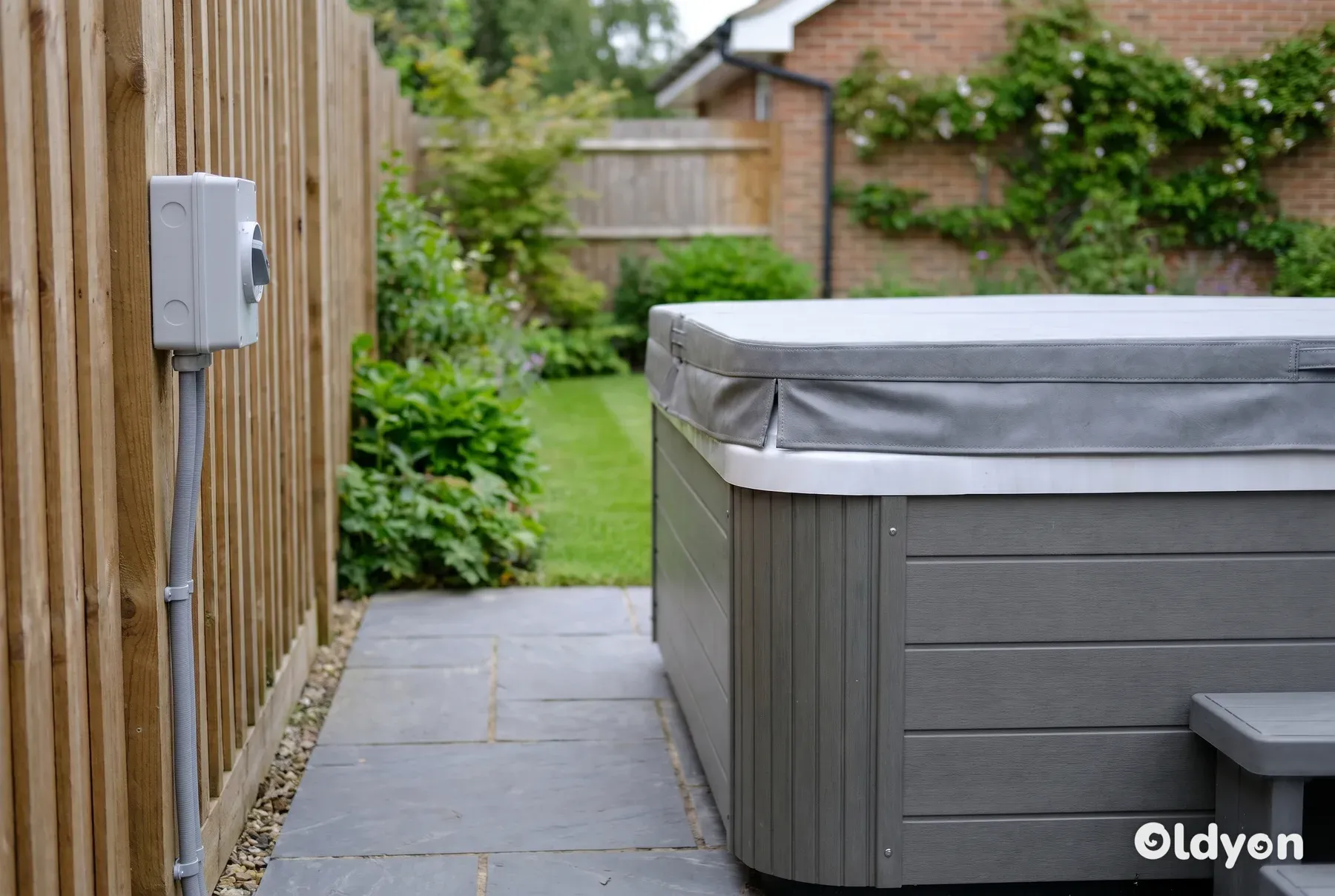Hartschalen-Whirlpool auf einer gepflasterten Terrasse in einem britischen Garten mit einem grauen, wetterfesten Trennschalter an einem nahegelegenen Holzzaun
