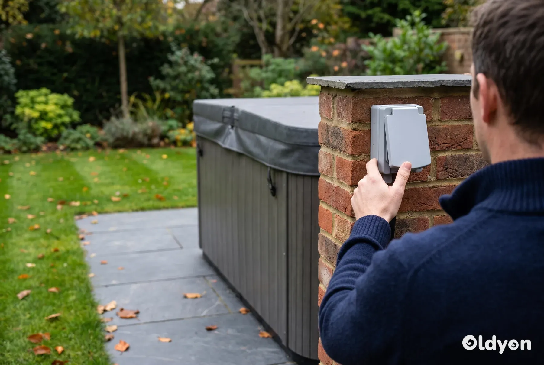 Person seen from behind switching off an outdoor isolator on a garden wall beside a covered hot tub on a stone patio