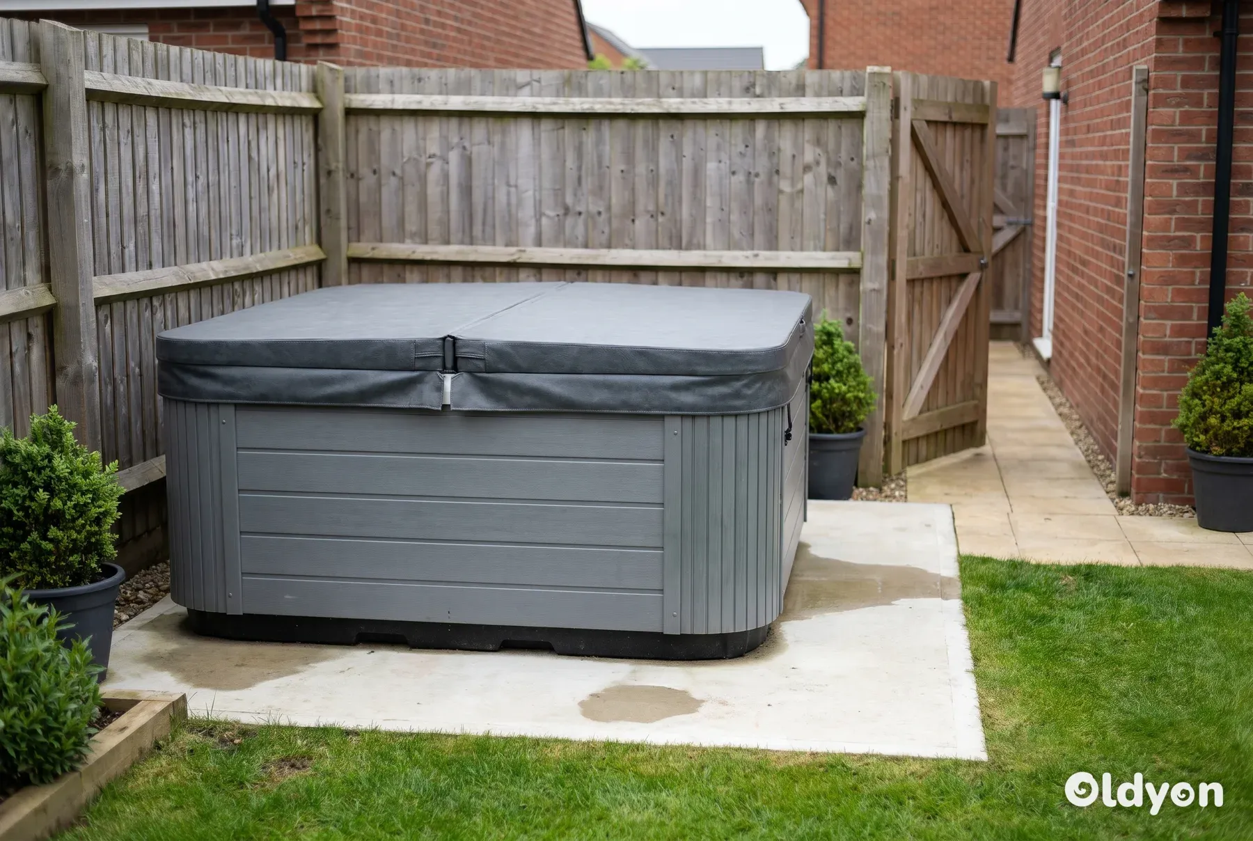 Un spa carré gris avec sa couverture fermée sur une dalle en béton à côté d'une clôture, avec un portail latéral en bois ouvert à proximité