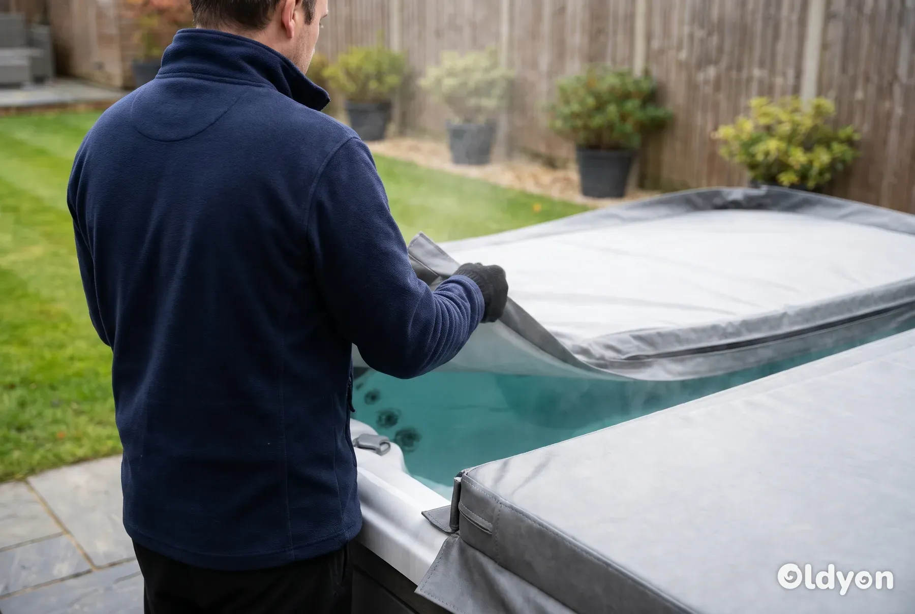 Persona vista de espaldas doblando hacia atrás la cubierta gris de una bañera de hidromasaje en un jardín británico, con un ligero vapor elevándose del agua cristalina