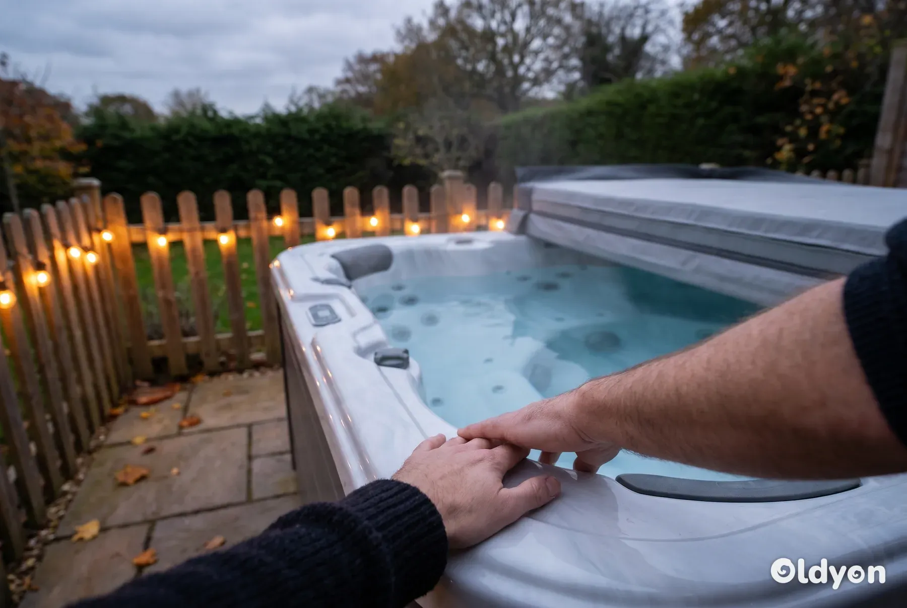 Steaming hot tub with its cover folded back in a British garden at dusk, one hand resting on the warm edge