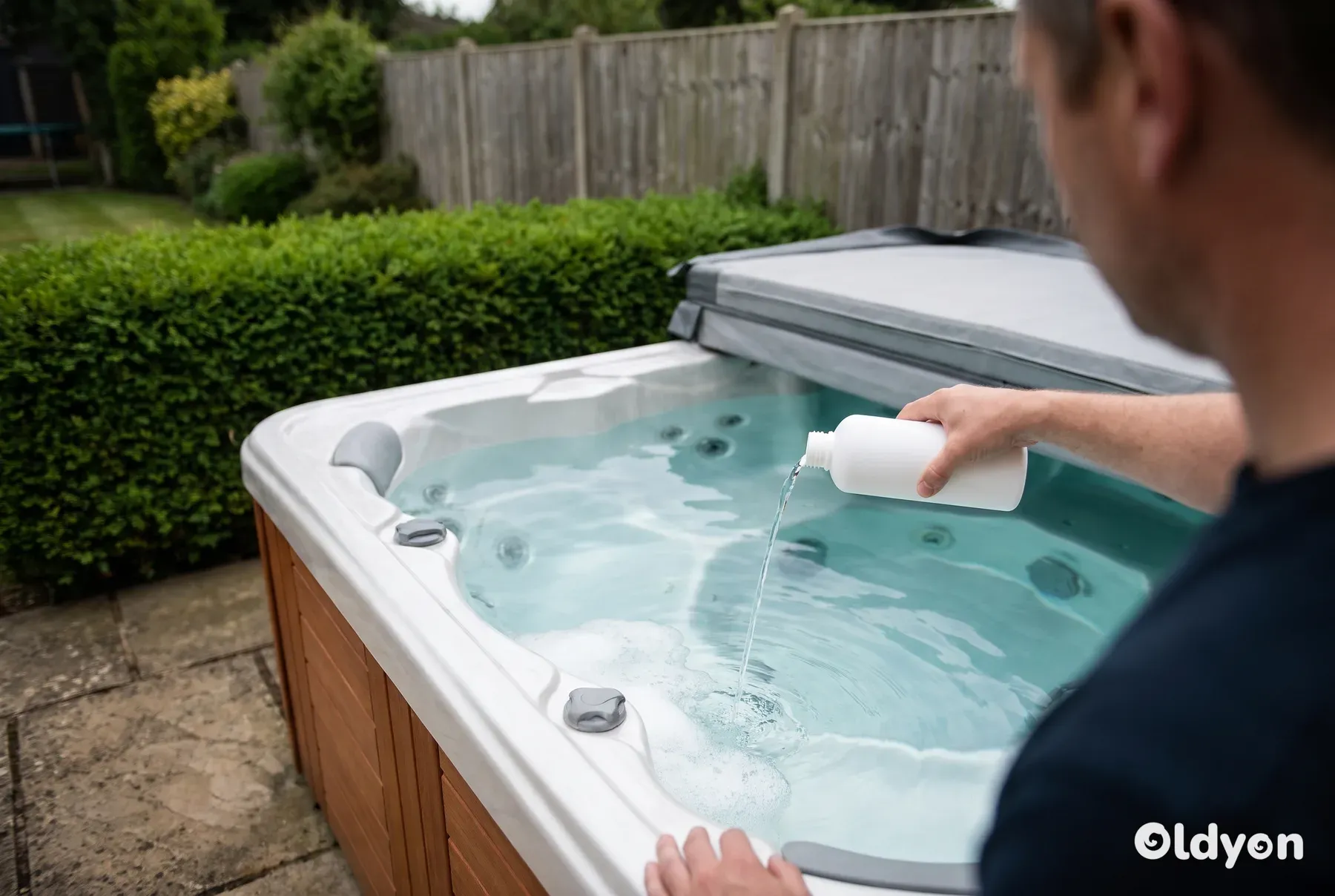 Eine Hand gießt eine kleine Dosis aus einer schlichten weißen Flasche in einen Whirlpool mit leichtem Oberflächenschaum in einem britischen Garten
