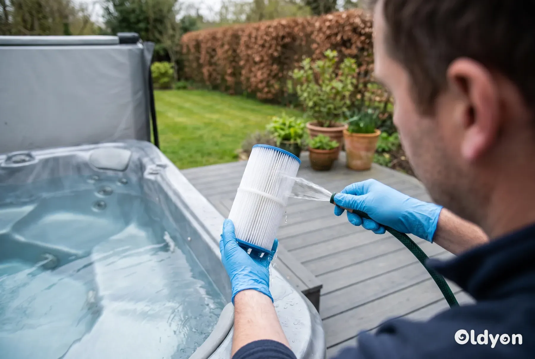 Hände mit Handschuhen spülen eine Whirlpool-Filterkartusche mit einem Gartenschlauch neben einem Whirlpool in einem britischen Garten ab