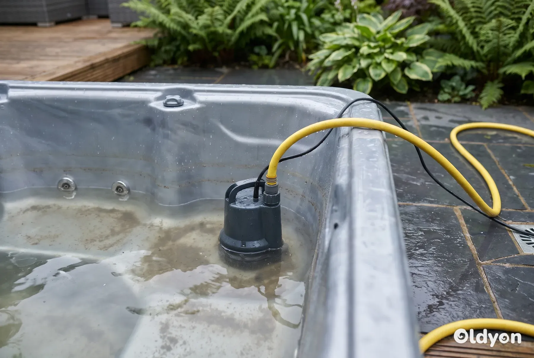 Una pompa sommersa collegata a un tubo da giardino giallo drena l'acqua bassa e torbida da una vasca idromassaggio.