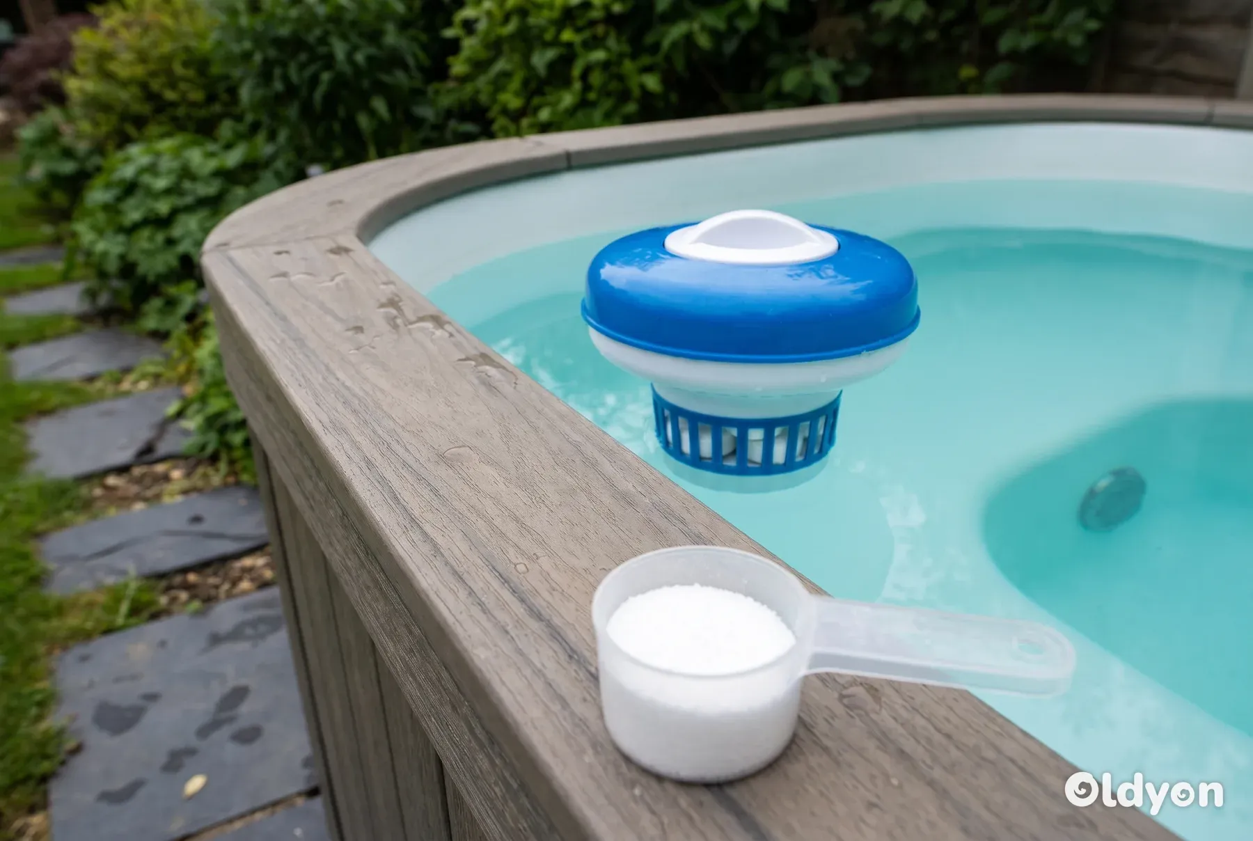 En flydende dispenser vugger i vandet ved siden af en skefuld hvidt granulat på trækanten.