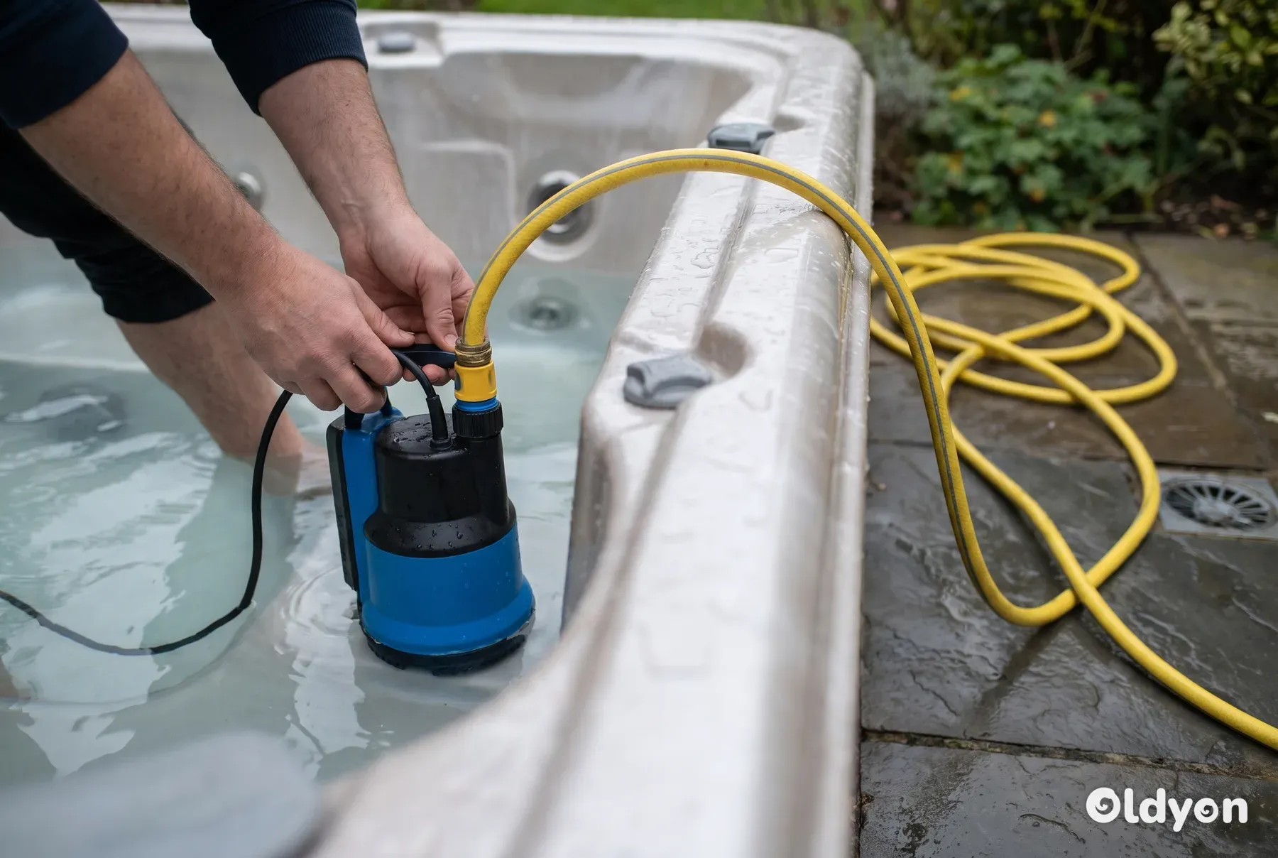 Een dompelpomp met een gele slang staat in een gedeeltelijk geleegde hot tub op een terras.