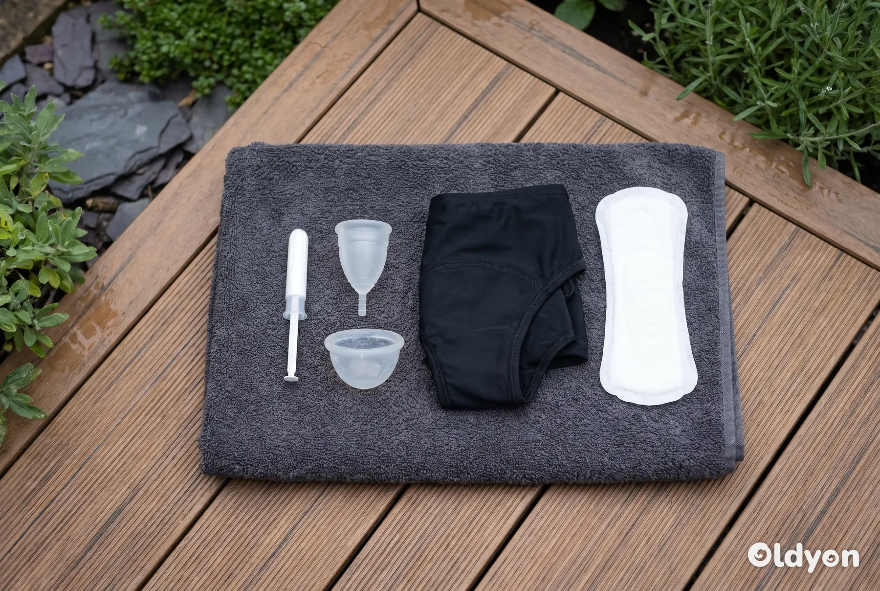 Vari prodotti mestruali senza marchio, tra cui una coppetta, un assorbente interno, un disco, un costume da bagno e un assorbente, disposti su un asciugamano.