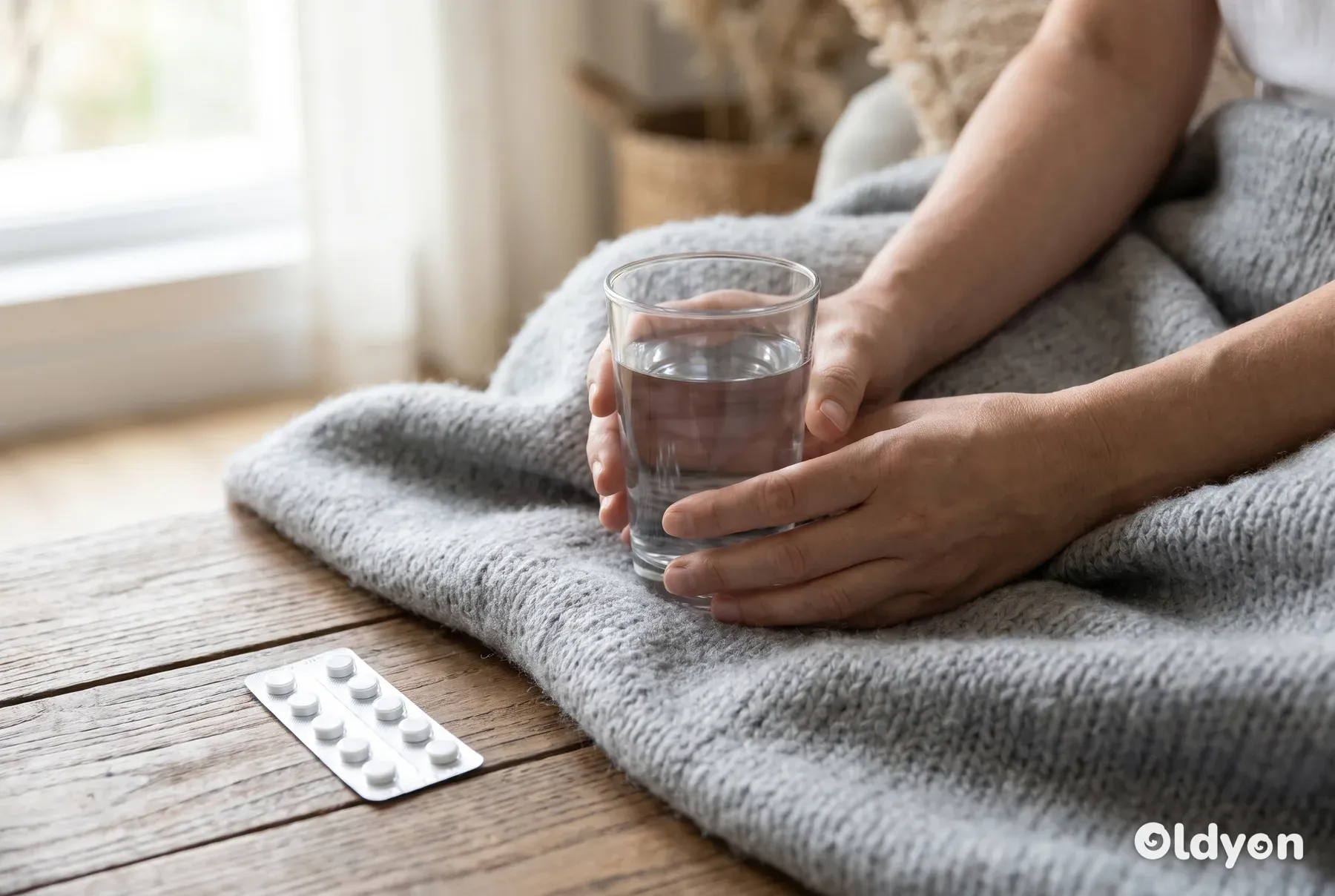 Hænder, der hviler på et blødt tæppe, holder et gennemsigtigt glas vand ved siden af medicin uden etiket.