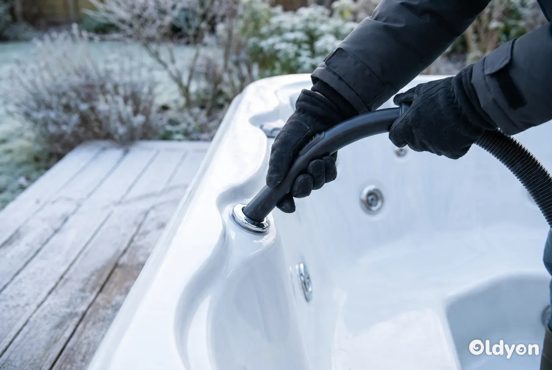 Hænder i handsker holder slangen fra en våd- og tørstøvsuger mod en dyse i et spabad i en frostklar have.